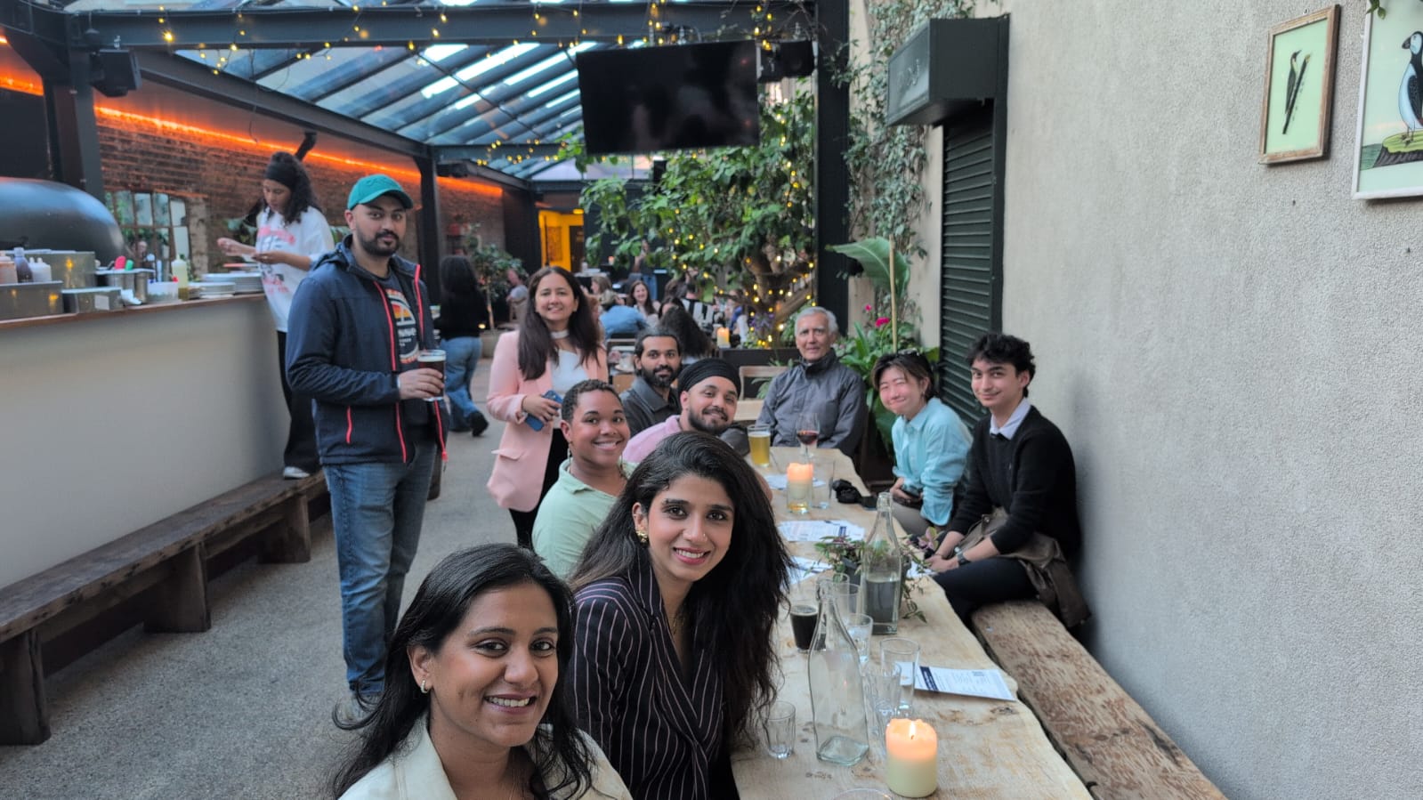 Selfie at the eChai AI Social in London at The Hawk's Nest: founders and operators seated around a long wooden table on a fairy-lit covered patio with hanging plants and brick walls