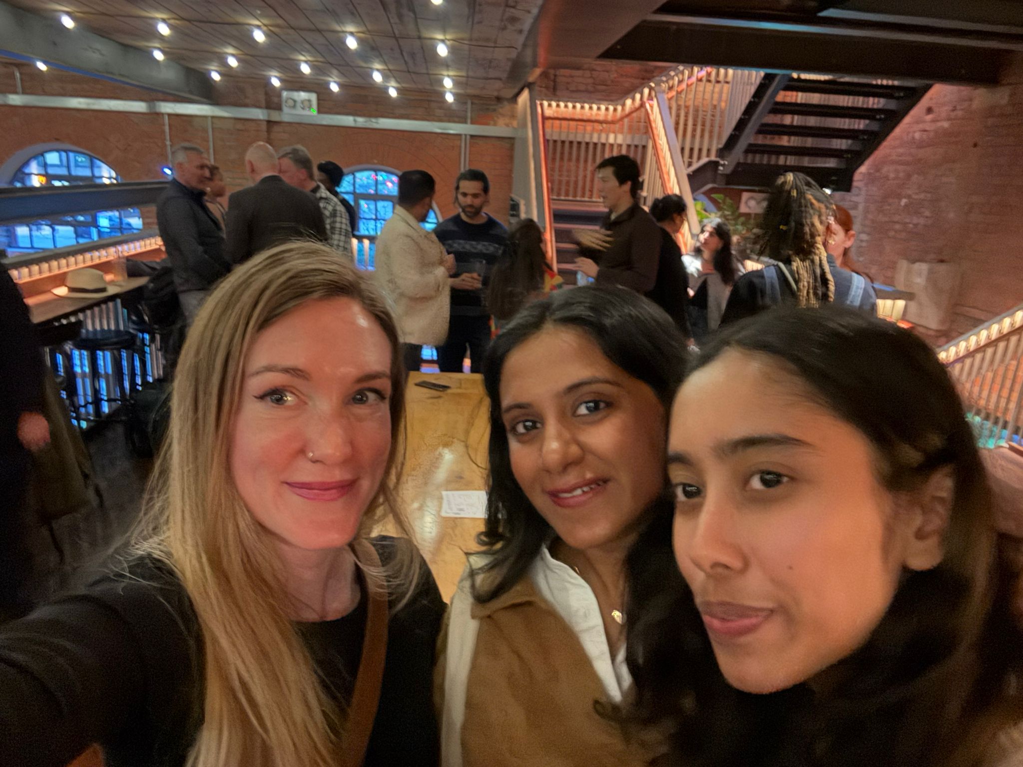 Selfie at the eChai London Social inside The Light Bar & Dining: three attendees in the foreground, a warmly lit exposed-brick room with festoon lights and other founders gathered behind them