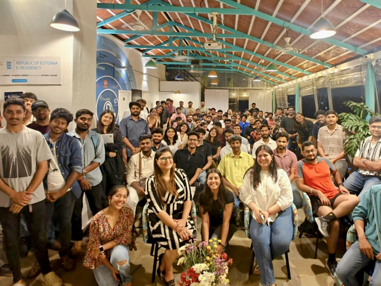 Packed group photo at an eChai Bengaluru meetup: about 80 early-stage founders gathered indoors under exposed trusses and warm pendant lights, with banners on the wall behind them