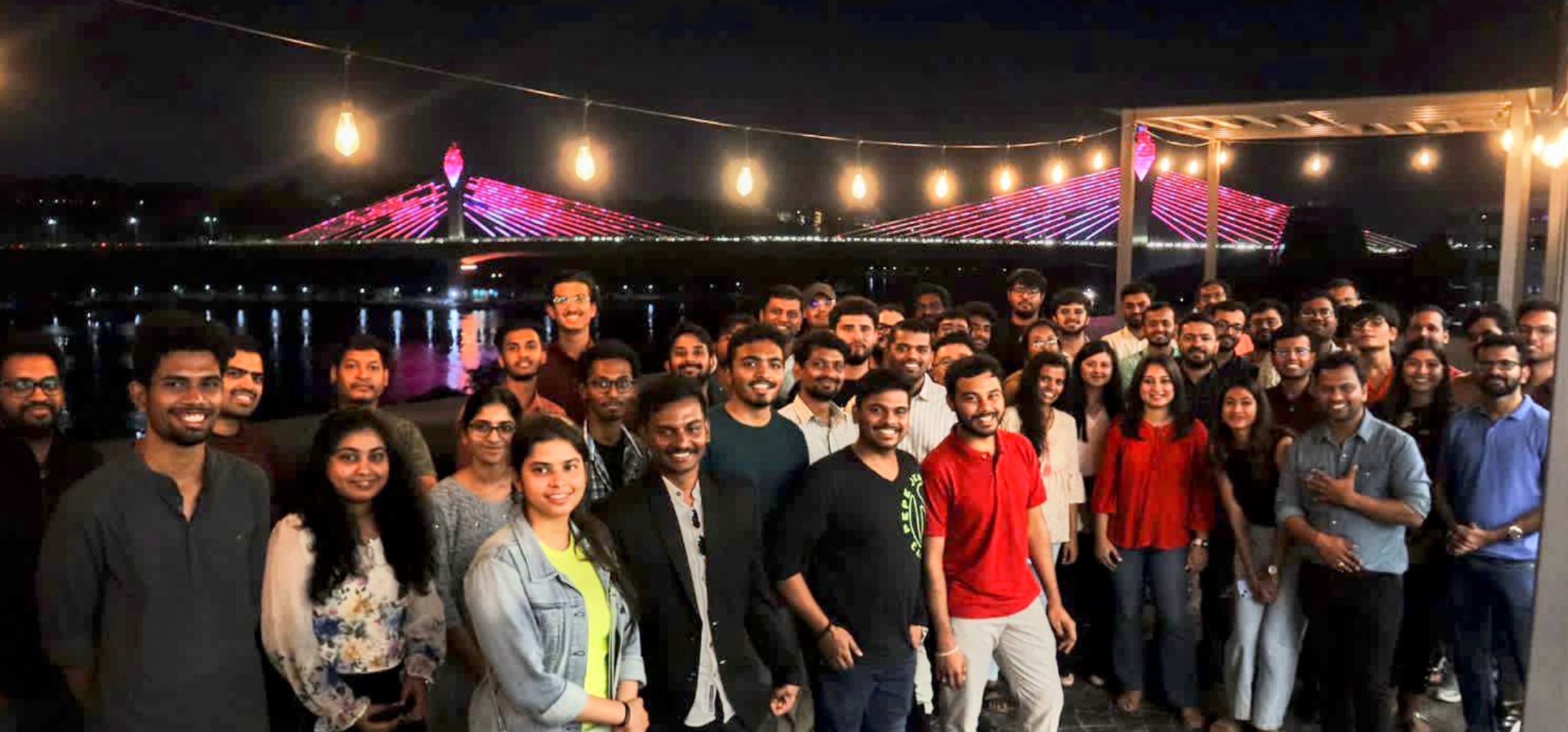 Group photo at an eChai Hyderabad meetup on a riverside rooftop: 60+ early-stage founders standing under string lights with the illuminated Durgam Cheruvu Cable Bridge in the background