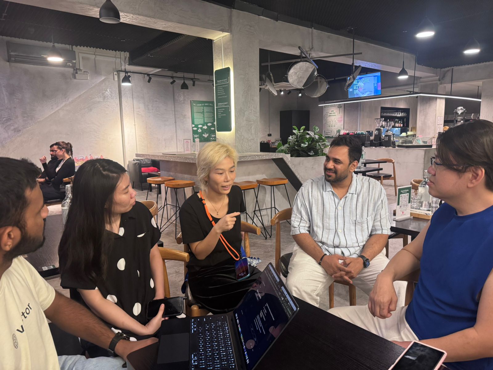 Small group of founders in conversation at Monk's Brew Club in Singapore: five people seated around a table with a laptop open, an industrial concrete cafe interior with a 'NETWORK WITH US' screen behind them