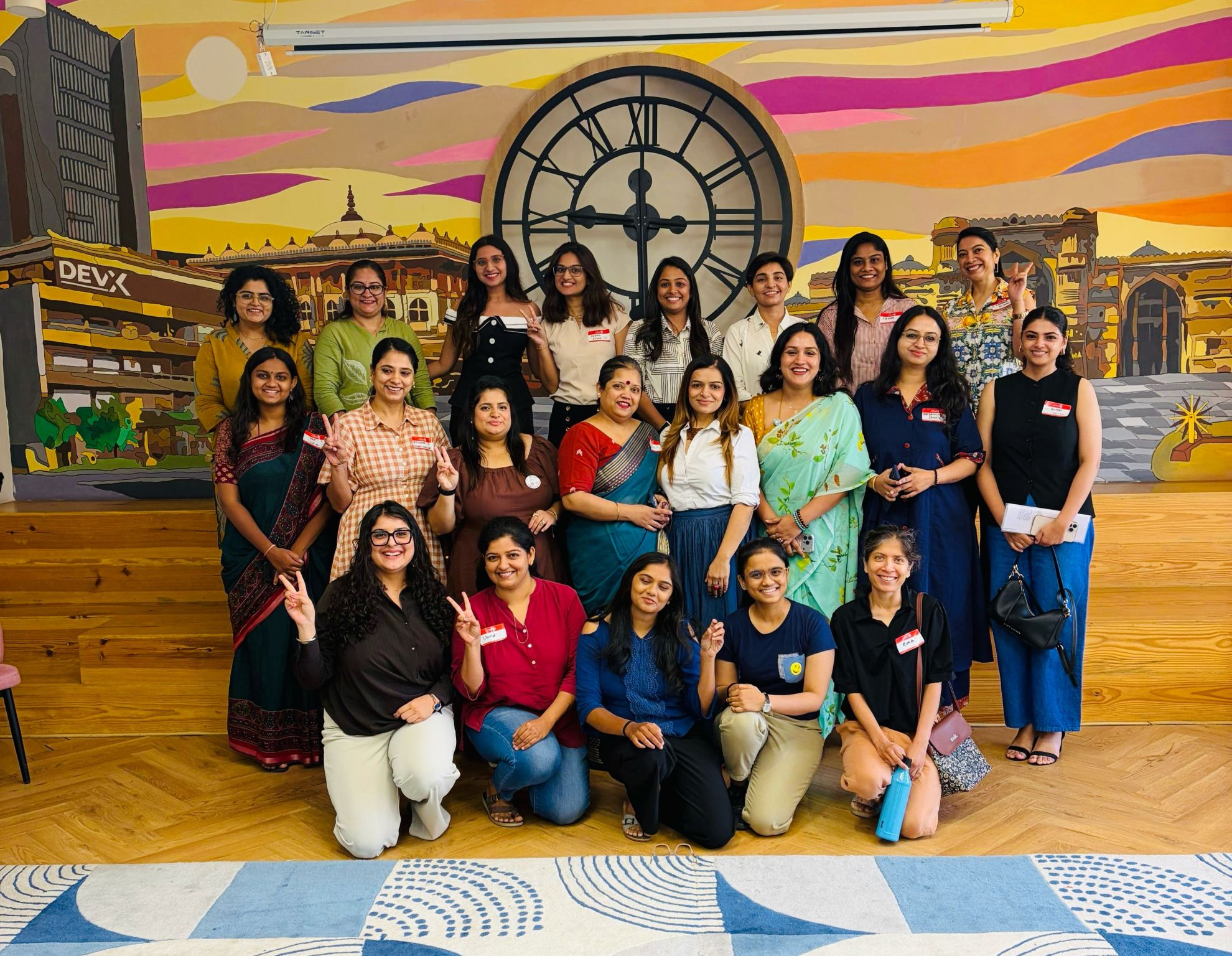 Group photo of the Women Founders Circle in Ahmedabad at DevX Sindhu Bhavan Road, women leaders standing together in front of a vivid mural of Ahmedabad landmarks under a sunset sky and a large clock