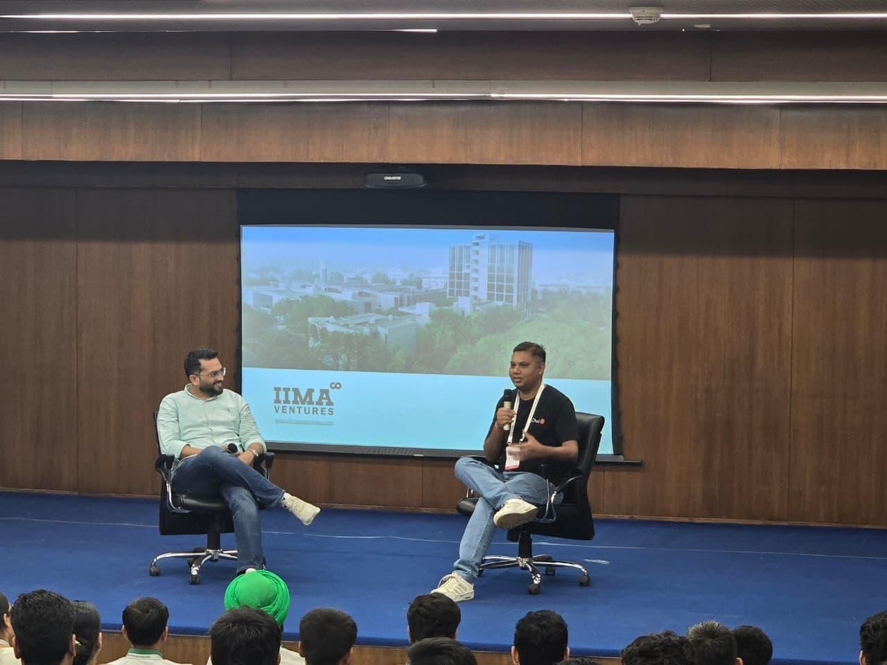 Jatin Chaudhary (eChai Ventures) on stage at IIM Ahmedabad moderating a fireside chat with Vishal Virani (Founder, Rocket); IIMA Ventures slide behind them showing the IIMA campus, with audience seated in the foreground