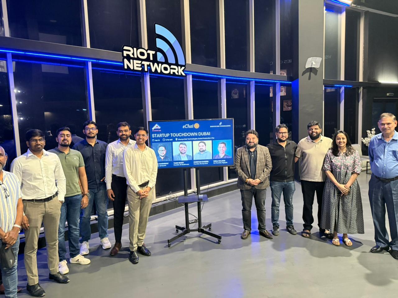 Group photo at the eChai Founders Social in Dubai: founders standing under the RIOT Network signage at Shunya Ekai HQ, with a 'Startup Touchdown Dubai' panel slide on the screen behind them