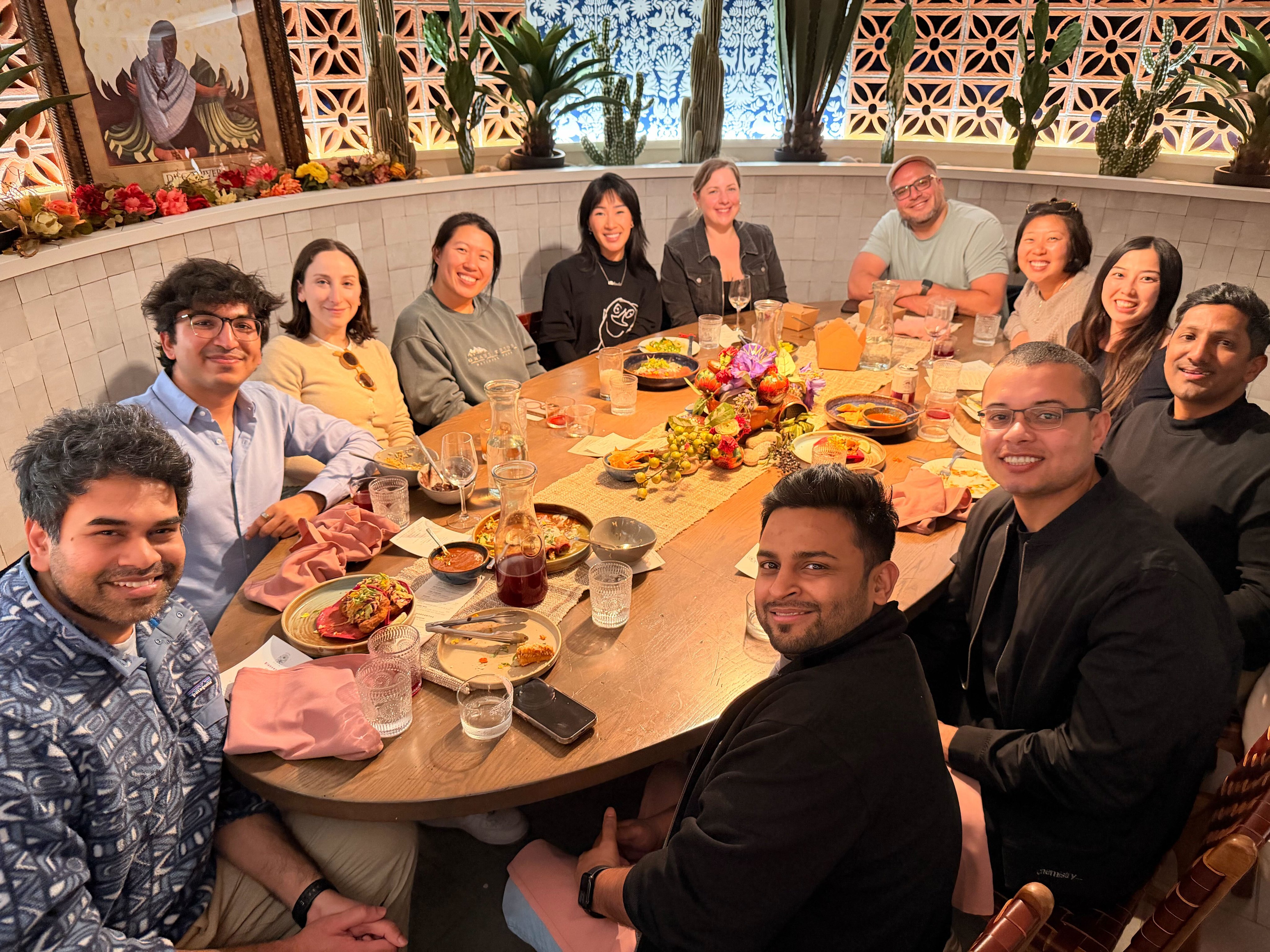 The Noon team gathered around a long oval table at a San Francisco restaurant, eleven people sharing a meal in front of a tiled wall and a row of cacti and floral arrangements